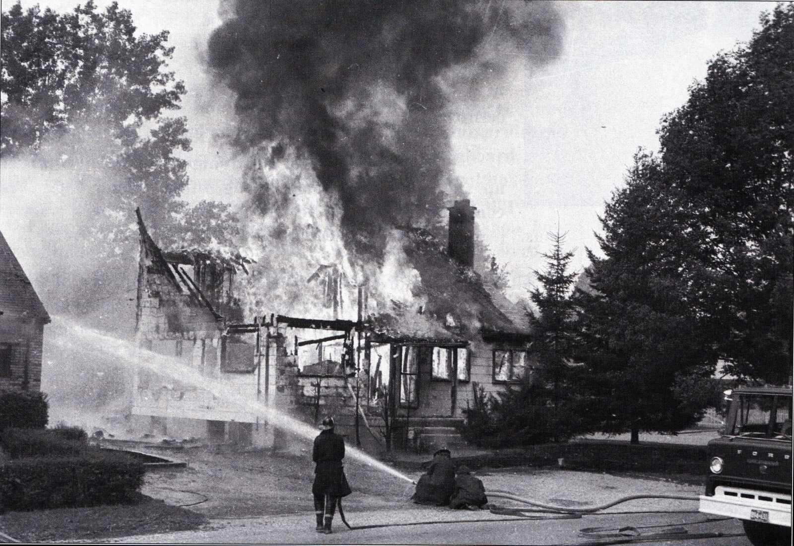 A black and white photo shows a house full engulfed in flames with a fire fighter holding a hose spraying water.